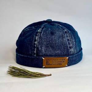 Puede incluir: Gorra de mezclilla azul oscuro con una parte superior pequeña y redonda. Un parche de cuero marrón con el texto "KARTCEV LEATHER GOODS" está cosido en la parte delantera. Una ramita de agujas de pino verdes descansa frente a la gorra.