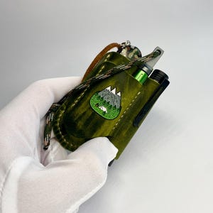 May include: A green leather case with a mountain-themed patch, holding a green vape pen. The case has a black clip and a camouflage lanyard. The leather has a distressed finish.