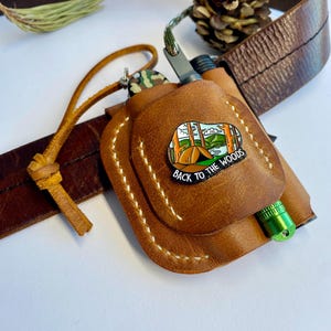 Puede incluir: Una funda de cuero marrón con un encendedor verde y un cuchillo pequeño. La funda tiene un diseño cosido y un pin que dice "Back to the Woods" con una escena de campamento. Una correa de cuero marrón y una piña están en el fondo.