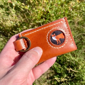 Op de afbeelding: Een bruine leren sleutelhouder met een gestikte rand en een cirkelvormige patch met een vosillustratie. De sleutelhouder heeft een metalen ring en een leren riem. De achtergrond is wazig groen gras.
