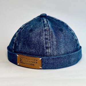 Op de afbeelding: Een donkerblauwe denim cap met een afgeronde kroon en een korte rand. Een bruin leren label met de tekst "KARTCEV LEATHER GOODS" is op de voorkant genaaid. De cap is een klassieke stijl, geschikt voor casual wear.