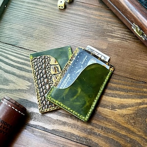 May include: Two leather card holders, one featuring a textured brown crocodile-embossed pattern and the other a smooth olive green finish with visible stitching. A bank card with the text KRS IBBANK is inserted into the green card holder.