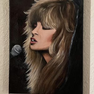 May include: An oil painting on canvas depicts a woman with long, layered blonde hair and closed eyes, singing into a microphone. The portrait features a dark background, with the woman's face and hair illuminated. The painting is mounted on a white wall.
