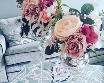 May include: Two silver glass vases with a floral arrangement of pink, peach, and white flowers. The flowers are arranged in a cascading style and are accented with green leaves.