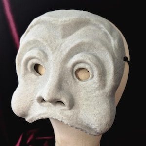 May include: A pale grey theatrical mask with a stylized face, featuring large eye openings and a prominent nose. The mask is mounted on a white stand against a dark background, suggesting a costume or prop.