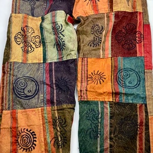 May include: Patchwork trousers in shades of orange, green, and brown. The design incorporates various geometric and sun motifs. The fabric looks to be a soft, lightweight material, suitable for casual wear.