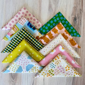 May include: A collection of colorful, triangular fabric pieces with various patterns. Designs include smiley faces, bears, stars, umbrellas, and floral prints. The fabrics are arranged on a light wooden surface.