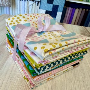 May include: A stack of colorful fabric squares tied with a light pink ribbon. The fabrics feature various patterns, including polka dots, smiley faces, and geometric designs. The color palette includes pink, green, blue, and yellow. The fabrics are likely for quilting or other craft projects.
