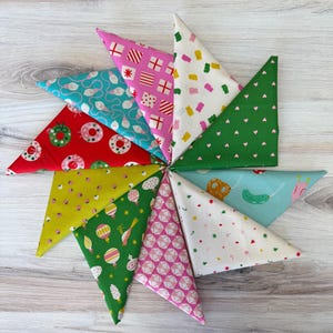 May include: A collection of ten triangular fabric swatches arranged in a circular fan pattern. The fabrics feature various colorful prints including holiday ornaments, gift boxes, gummy bears, small hearts, pretzels, and geometric patterns.