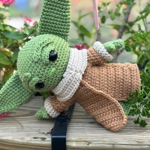 May include: A crocheted green and brown plush toy resembling a character from the Star Wars franchise. The toy is wearing a brown coat and a white scarf.