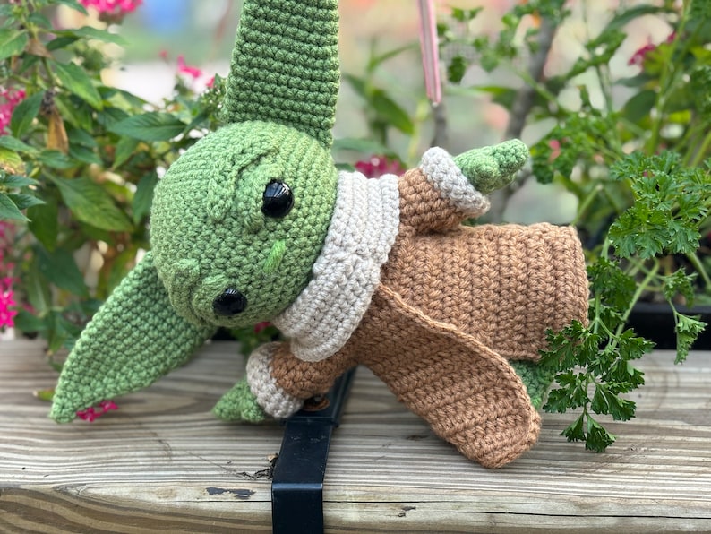 May include: A crocheted green and brown plush toy resembling a character from the Star Wars franchise. The toy is wearing a brown coat and a white scarf.