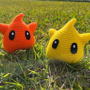 May include: Two handmade crochet star-shaped plush toys. One is orange, the other yellow. Both have black oval eyes with white highlights. The toys are sitting on green grass in natural sunlight. The stars have a whimsical, cartoonish design.