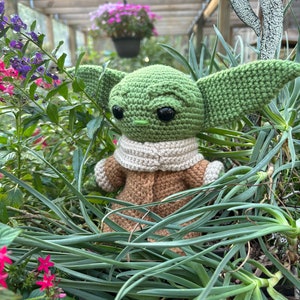 May include: A green crocheted plush toy resembling a character from the Star Wars franchise, with large eyes and a brown crocheted coat, is nestled in a bed of green plants.