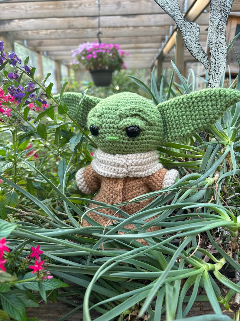 May include: A green crocheted plush toy resembling a character from the Star Wars franchise, with large eyes and a brown crocheted coat, is nestled in a bed of green plants.