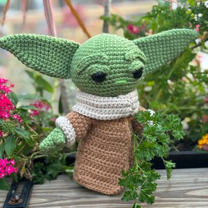 May include: A green crocheted Yoda doll wearing a brown crocheted coat and a white crocheted scarf. The doll is holding a sprig of green parsley.