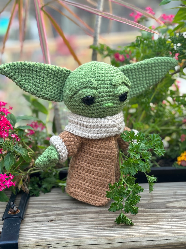 May include: A green crocheted Yoda doll wearing a brown crocheted coat and a white crocheted scarf. The doll is holding a sprig of green parsley.