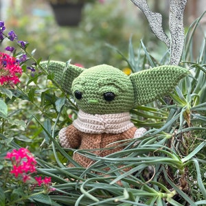 May include: A green crocheted plush toy with large eyes and a brown crocheted coat. The toy is sitting in a bed of green plants.