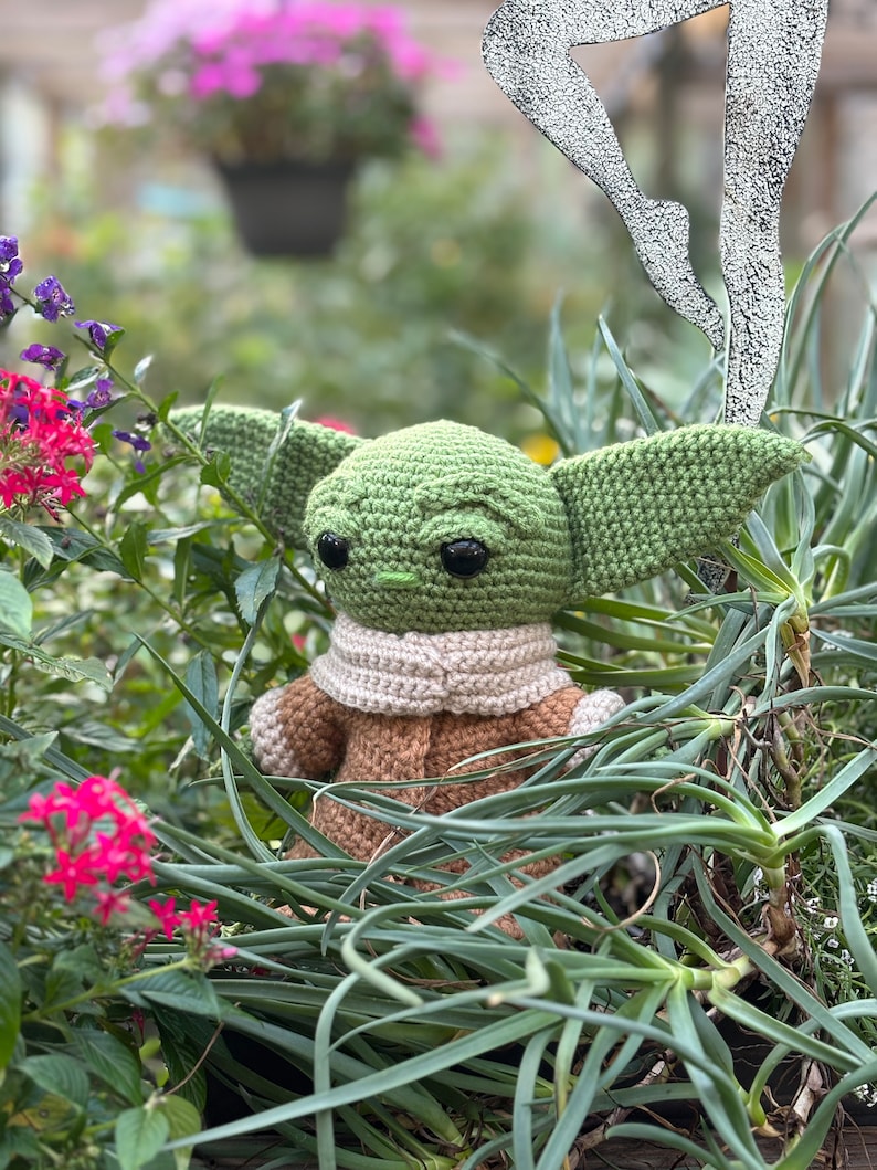 May include: A green crocheted plush toy with large eyes and a brown crocheted coat. The toy is sitting in a bed of green plants.