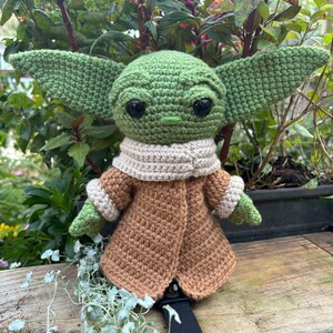 May include: A green crocheted Yoda doll wearing a brown coat with a cream colored scarf. The doll is sitting on a wooden surface.