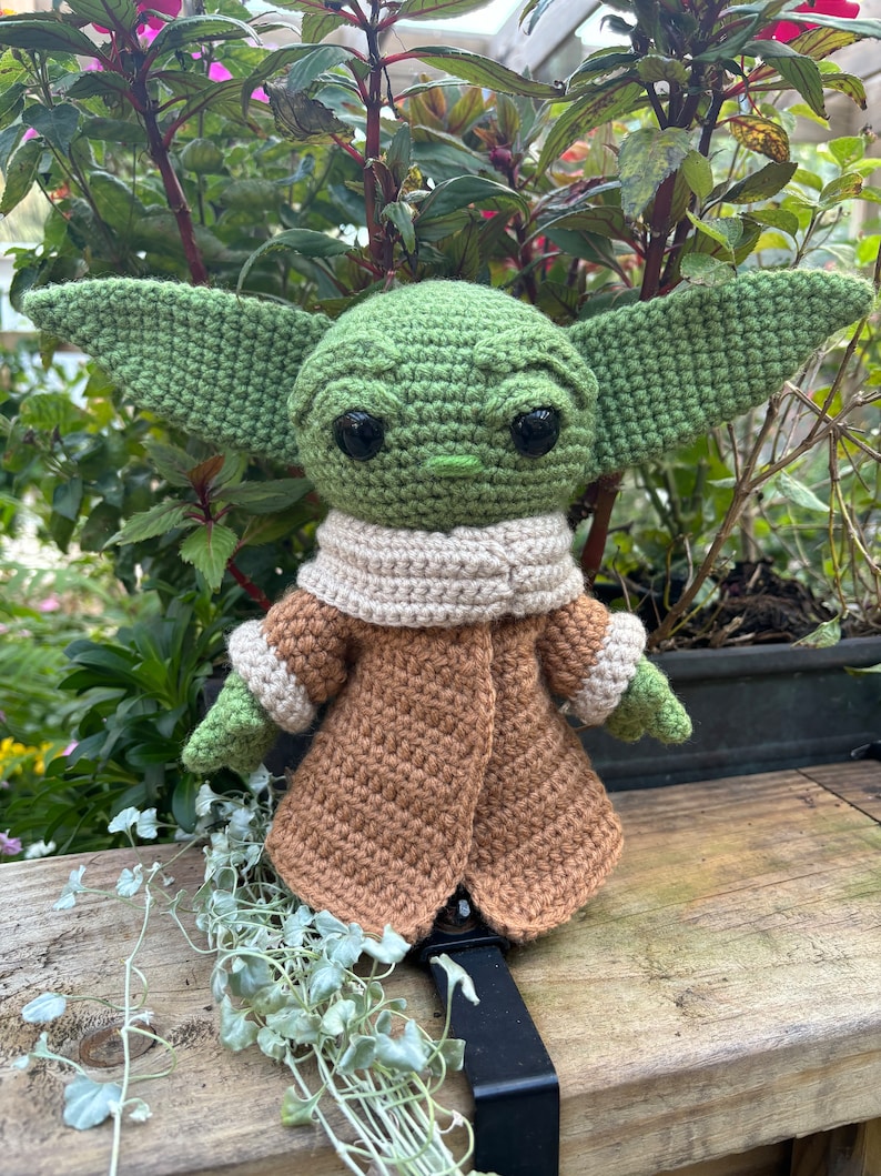 May include: A green crocheted Yoda doll wearing a brown coat with a cream colored scarf. The doll is sitting on a wooden surface.