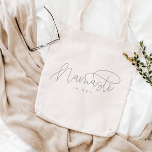 May include: A white canvas tote bag with the text "Namaste in Bed" in black cursive script. The bag is lying on a white and beige blanket with a sprig of green foliage.