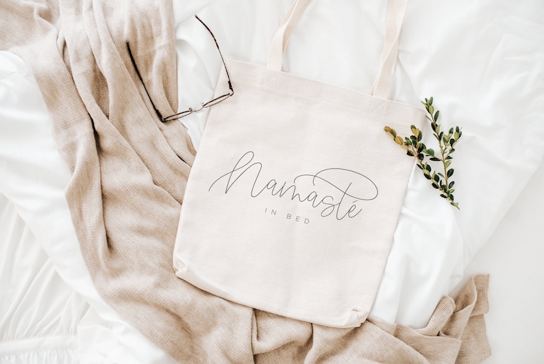 May include: A white canvas tote bag with the text "Namaste in Bed" in black cursive script. The bag is lying on a white and beige blanket with a sprig of green foliage.