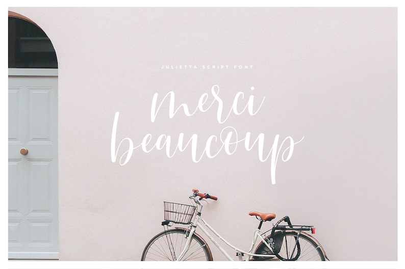May include: A white bicycle with a basket and a brown seat is parked in front of a light pink wall with the text "Merci beaucoup" written in white cursive script.