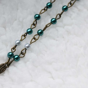 May include: A necklace with teal and silver beads on a bronze chain, with a bronze wing pendant. The necklace is displayed on a white, textured surface. The chain is made of a series of small links.
