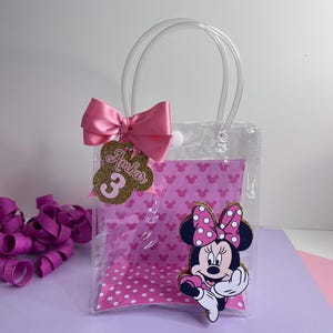 Puede incluir: Bolsa de regalo transparente con un lazo rosa y un gr&aacute;fico de Minnie Mouse. La bolsa presenta un interior de lunares rosas y una etiqueta dorada con el nombre "Ambar" y el n&uacute;mero "3". La bolsa tiene un asa transparente.