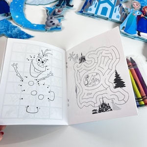 May include: An open activity book featuring a dot-to-dot drawing of a snowman on the left page and a maze with character illustrations on the right page. A small clear plastic case containing four colored crayons sits to the right of the book.