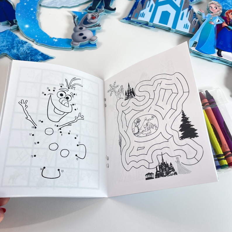 May include: An open activity book featuring a dot-to-dot drawing of a snowman on the left page and a maze with character illustrations on the right page. A small clear plastic case containing four colored crayons sits to the right of the book.