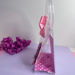 Puede incluir: Una bolsa de mano de pl&aacute;stico transparente con un interior de lunares rosas y un gran lazo rosa. La bolsa tiene asas transparentes y est&aacute; parcialmente llena de art&iacute;culos rosas. La cinta rizada morada est&aacute; esparcida sobre la superficie.