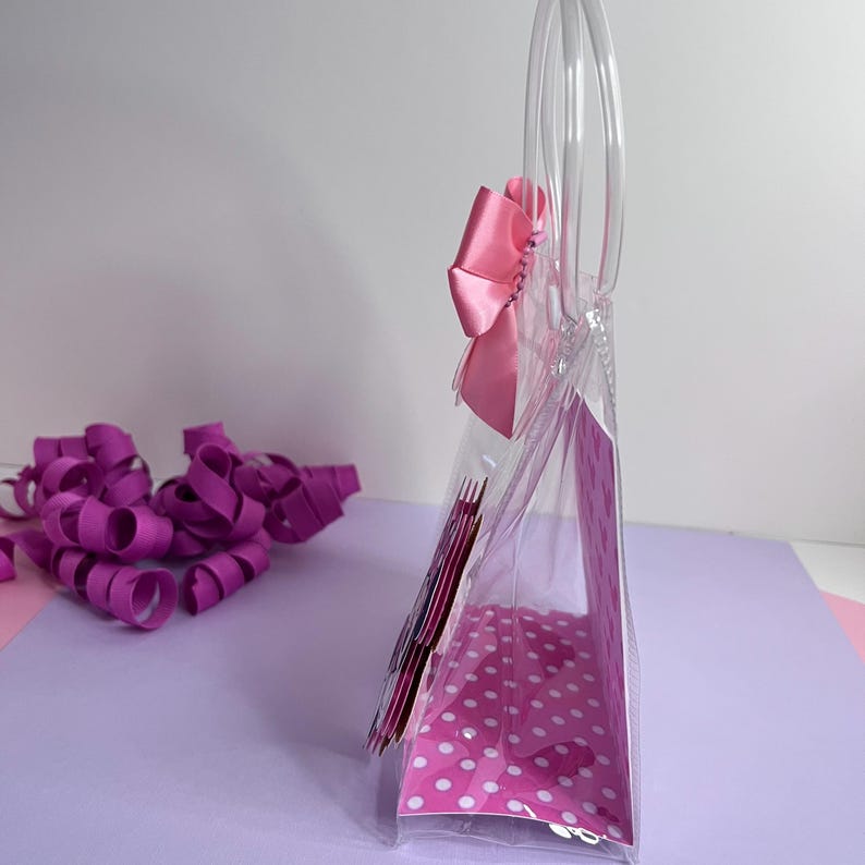 Puede incluir: Una bolsa de mano de pl&aacute;stico transparente con un interior de lunares rosas y un gran lazo rosa. La bolsa tiene asas transparentes y est&aacute; parcialmente llena de art&iacute;culos rosas. La cinta rizada morada est&aacute; esparcida sobre la superficie.