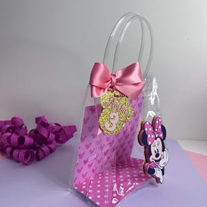Puede incluir: Una bolsa de regalo de pl&aacute;stico transparente con un lazo rosa y una etiqueta dorada con purpurina que dice "Ambar 3". La bolsa tiene un estampado de lunares rosas y Mickey Mouse. Un recorte de Minnie Mouse est&aacute; adjunto.