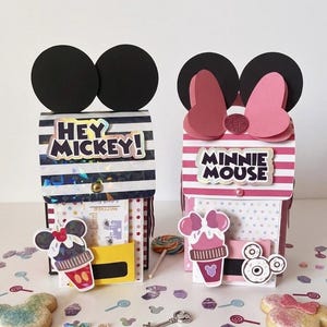 Könnte beinhalten: Zwei dekorative Leckerli-Boxen aus Papier mit charakterinspirierten Designs. Die linke Box hat schwarz-weiße Streifen mit dem Text HEY MICKEY! und einer Mickey-Eiscreme-Grafik. Die rechte Box hat rosa-weiße Streifen mit einer Schleife, dem Text MINNIE MOUSE und einer Minnie-Eiscreme-Grafik.