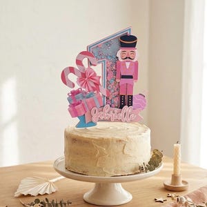 Op de afbeelding: Een cake met glazuur op een witte taartstandaard, versierd met een taarttopper. De topper bevat een roze notenkraker, zuurstokken, cadeauverpakkingen en de naam "Gabrielle". Rechts staat een brandende kaars.