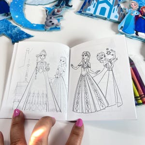 May include: A small coloring book featuring black and white line art illustrations of two female characters in long gowns on the left page and two female characters in detailed dresses on the right page, with a set of crayons visible on the side.