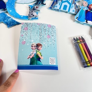 May include: A rectangular coloring book featuring Elsa and Anna on the cover with a light blue background, silver glitter accents, and pink snowflakes. A small QR code is printed on the bottom right. A clear plastic bag contains four crayons.