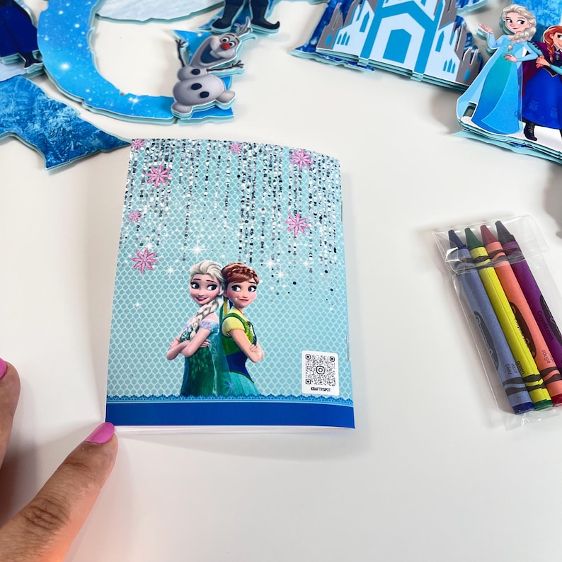 May include: A rectangular coloring book featuring Elsa and Anna on the cover with a light blue background, silver glitter accents, and pink snowflakes. A small QR code is printed on the bottom right. A clear plastic bag contains four crayons.