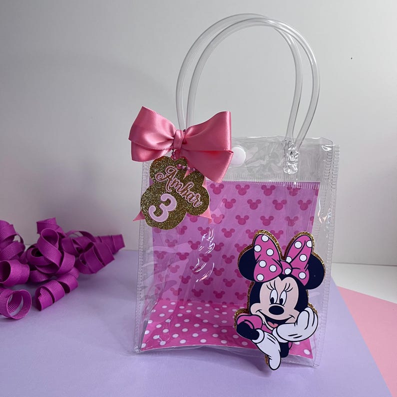 Puede incluir: Una bolsa de regalo de pl&aacute;stico transparente con un lazo y asa rosas. La bolsa presenta un gr&aacute;fico de Minnie Mouse y una etiqueta con el nombre "Ambar" y el n&uacute;mero "3". El interior tiene un estampado de lunares rosas.