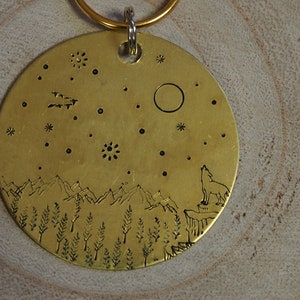 Op de afbeelding: Een sleutelhanger van messing met een ontwerp van een wolf die huilt naar de maan, bergen en bomen. Het ontwerp is geëtst in het messing en heeft een rustieke uitstraling.