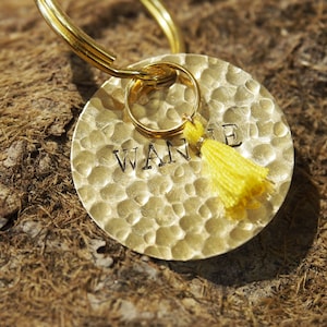 Op de afbeelding: Een gouden metalen sleutelhanger met een rond label waarop in zwarte letters "WANTE" staat. Het label heeft een gehamerde textuur en een gele kwast eraan bevestigd.