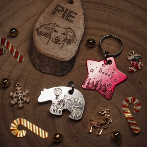 May include: A collection of personalised pet tags featuring various designs. A wooden tag with the name "PIE" and a dog illustration, a pink star-shaped tag with the name "Buddy" and a forest scene, a silver bear-shaped tag with the name "BUDDY" and a hot air balloon illustration, a gold reindeer charm, and several gold and red candy cane charms.