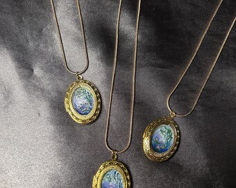 May include: Three gold-toned oval pendant necklaces. Each pendant features a miniature painting of a blue and purple floral scene. The necklaces hang on delicate gold chains, set against a dark, textured background.