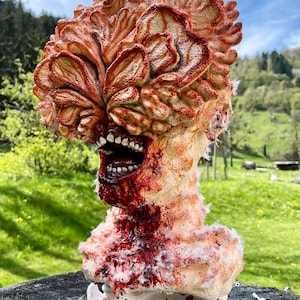 May include: A hand-painted, detailed sculpture of a monstrous creature with a fleshy, brain-like head, a gaping mouth with teeth, and a bloody, fleshy neck. The sculpture is made of a white material and is sitting on a wooden surface.