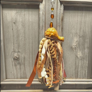 May include: A decorative hanging tassel ornament featuring a glazed ceramic turkey leg topper in a golden brown finish. The tassel is composed of various ribbons, including brown, tan, and white polka dot patterns, alongside textured fabric strips.