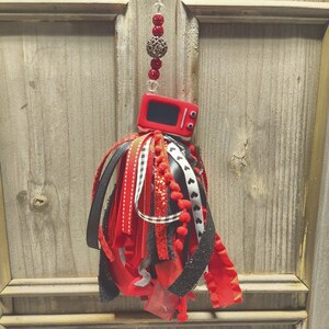 May include: A decorative hanging ornament featuring a miniature red television set with a black screen, suspended from a vertical string of red and clear beads. Below the television is a cluster of ribbons in red, black, and white patterns.