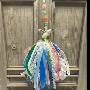 May include: A hanging ornament featuring a ceramic hummingbird figurine perched atop a sphere adorned with a dense collection of multi-colored ribbons, lace trims, and fabric strips in shades of blue, green, pink, and white, topped with beads.