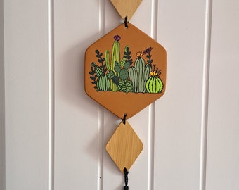 May include: A terracotta hexagon-shaped wall hanging with a painted design of cacti and succulents. The hanging is attached to a wooden diamond shape with a black tassel.