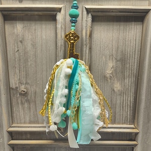 May include: A decorative tassel hanging from a gold-colored metal key ornament. The tassel consists of various ribbons, lace, and fabric strips in shades of white, turquoise, and gold, accented with small white pom-poms and a string of turquoise beads.